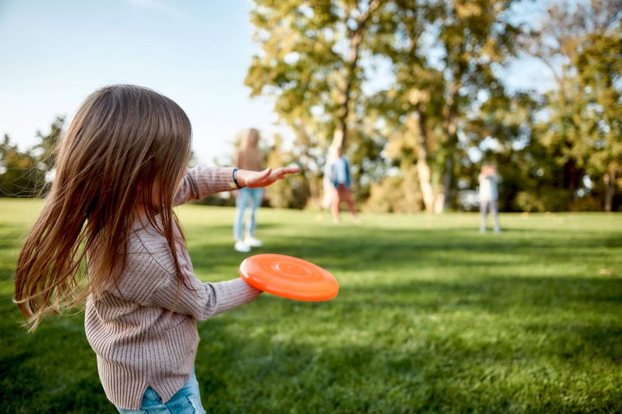 Attain at Harbour View luxury apartments with Young girl with long hair prepares to throw an orange frisbee in a sunny park with people in the background.