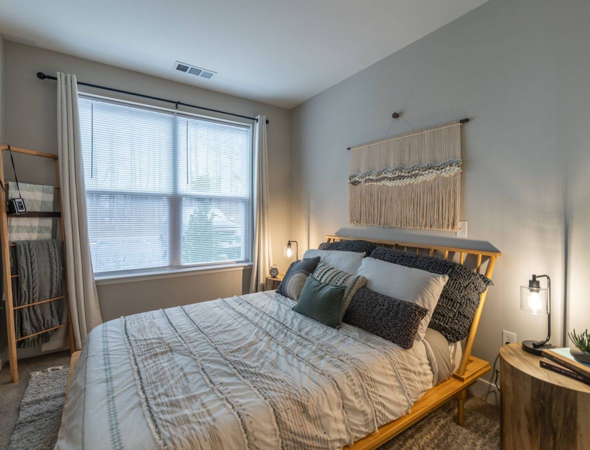 Attain at Harbour View luxury apartments with Cozy bedroom with a wooden bed, neutral bedding, wall art, a round nightstand, and large window with blinds.