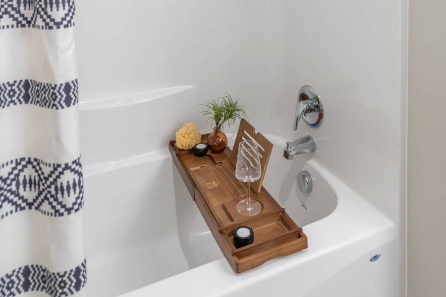 Attain at Harbour View luxury apartments with White bathtub with a shower, patterned curtain, and a wooden bath tray holding decor and a wine glass.